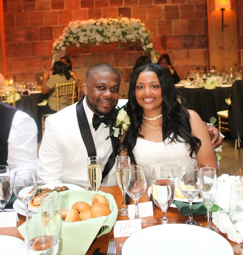 A bride and groom smiling at their head table with plates in front of them and a floral arch behind